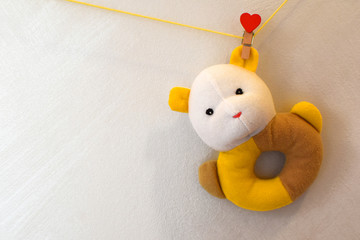 Little toy bear hanging on yellow rope with red heart shaped peg on light grey wall background with copy space for text. Soft rattle toy for newborn babies.