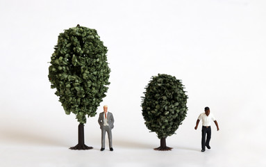 A miniature white man and a miniature black man standing next to a tree of different sizes.