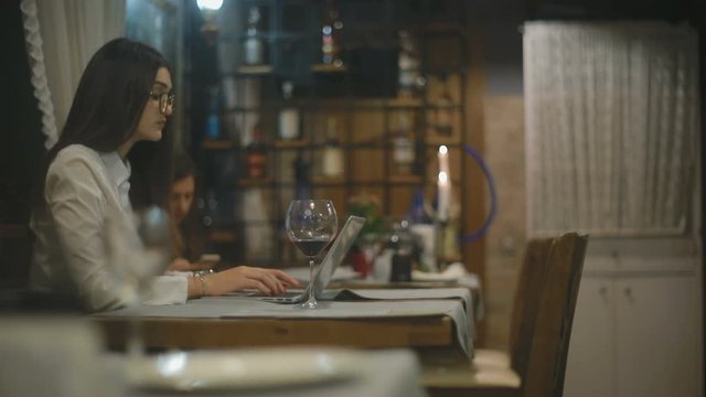 slow motion brunette young woman in glasses with loose flowing hair works on laptop and has sip of red wine in cafe