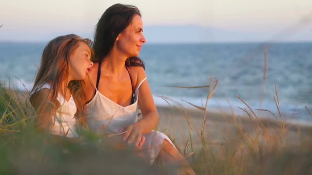 happy mother with her little daughter sitting on the beach at sunset, hugging and playing. concept of happy mothers and daughters