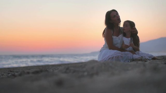 happy mom with her little daughter sitting on the beach at sunset, hugging and playing. concept of happy mothers and daughters