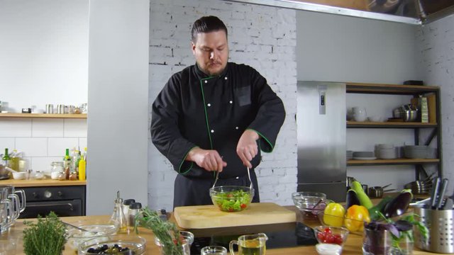 Tracking shot of male restaurant chef in uniform tossing salad in glass bowl. Fresh vegetables, herbs and olive oil are on table