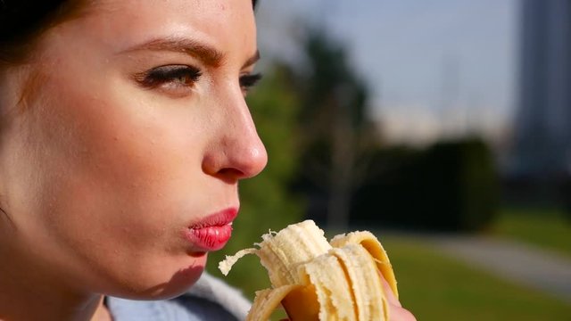 Slow motion, close-up attractive sexy young brunette woman with beautiful eyes and red lips eats banana on sunny day. Spring beriberi, vitamins in fruits.