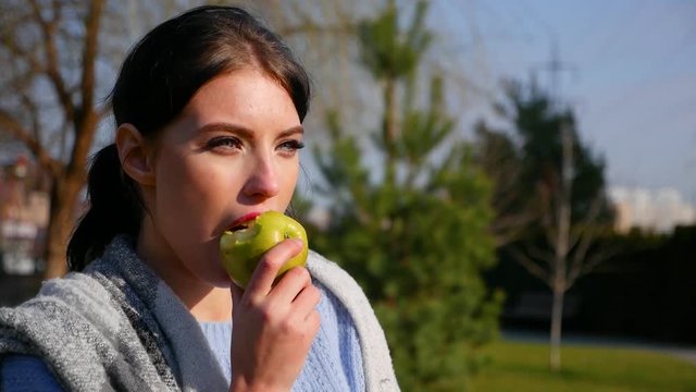 Slow motion, close-up attractive sexy young brunette woman with beautiful eyes and red lips eats green apple on sunny day. Spring beriberi, vitamins in fruits.