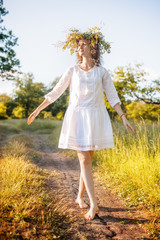 Caucasian happy girl in a white dress with a wreath of wild flowers in the summer at sunset in the field.