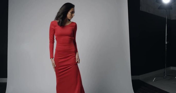 View over the shoulder of the photographer on the charming gorgeous young female model in the red long dress posing on the white background of the studio.
