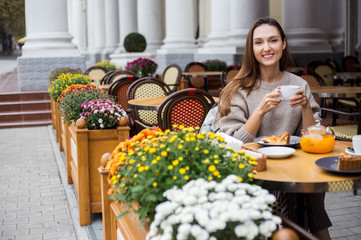 Young attractive smiling woman in gray knitted sweater drinking tea outdoors at the street cafe.