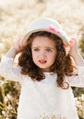 portrait of an upset  little girl with curly hair and with a straw hat in a daisy field