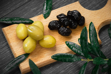 Green and black olives on the wooden plate with olive-tree leaves on black background.