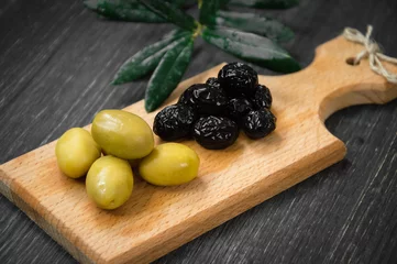 Foto op Plexiglas Green and black olives on the wooden plate with olive-tree leaves on black background. © Aydar