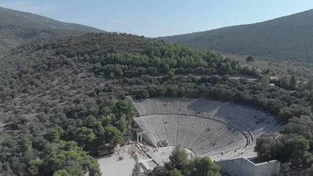 Panning from right to left and then clockwise showing the side and front of the ancient theater of Epidavros in Greece through forests and mountaints. Clear sky, sunny weather, flat colour profile.