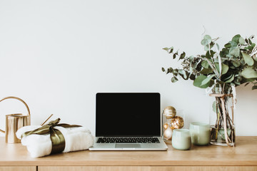 Blogger, freelance home office desk workspace. Laptop with mock up screen on wooden desktop decorated with eucalyptus bouquet, gift box, candles on white background.