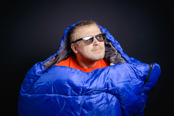 A young guy in sunglasses with a thoughtful look posing in a sleeping bag on a black background. The concept of tourism, camping.