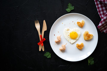 Breakfast on Valentine's Day fried eggs,sausage, and bread in the shape of a heart and fresh greens.Fork and knife made of ecological materials tied with a red ribbon.Top view.Valentine's Day concept