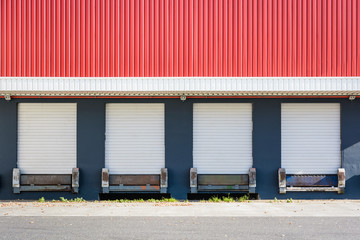 Front view of a truck loading dock at a warehouse in the suburbs of Paris, France, with four white roller shutter doors closed under a red corrugated metal siding.