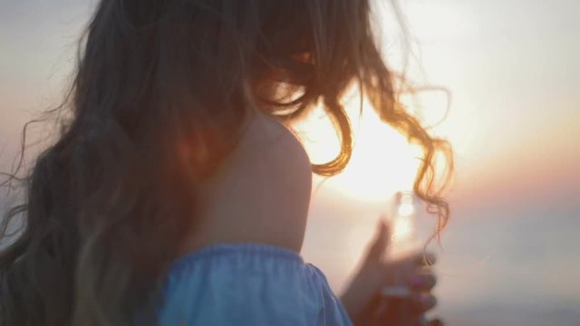 slow motion closeup blurry image girl in open shoulder summer dress with loose flowing hair holds bottle at sunset