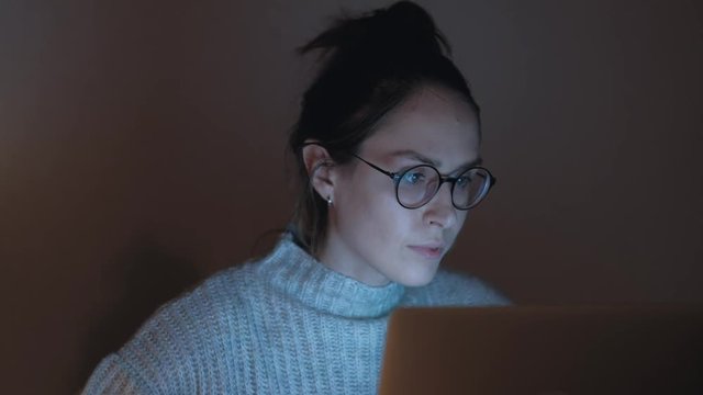 Cute woman in glasses browsing the Internet in dark room. 4k UHD. Portrait of a female working at the computer at night. 