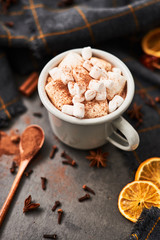Hot cocoa drink with marshmallows in retro mug surrounded by ingredients: cinnamon, orange, anis and cove on black concrete table. Winter drink. Top view. Copy space for text.