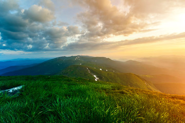 Wide summer mountain view at sunrise. Glowing orange sun raising in blue cloudy sky over green grassy hill soft grass and distant mountain range covered with morning mist. Beauty of nature concept.
