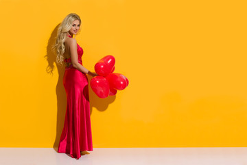 Obraz na płótnie Canvas Beautiful Woman In Elegant Red Dress Is Posing With Heart Shaped Balloons