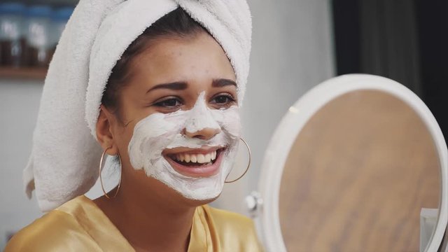 Emotional portrait of a happy and calm beautiful naked girl with a blue clay cosmetic mask on half of her face, looking at the camera mirror applying clay with a brush.