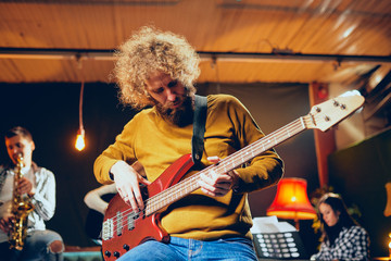 Close up of bass guitarist playing guitar while sitting on the chair. In background saxophonist and piano player. Home studio interior.