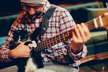 Caucasian musician with cap on head playing with cat while playing bass guitar.