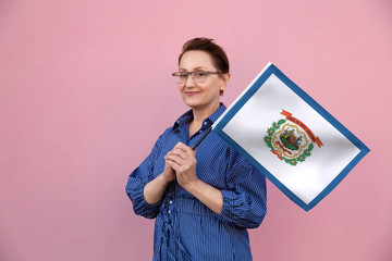 West Virginia flag. Woman holding West Virginia state flag. Nice portrait of middle aged lady 40 50 years old holding a large state flag over pink wall background on the street outdoor.