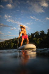 Handsome young man on a paddle board. Getting a great exercise on a lovely river in warm evening sunlight