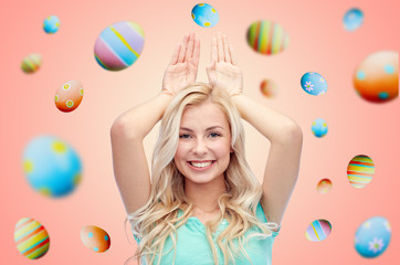easter, holidays and people concept - happy smiling young woman making bunny ears over pink or living coral background with colored eggs
