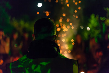 A man wearing a fluorescent jacket stares at a fire, with sparks and people visible in the...