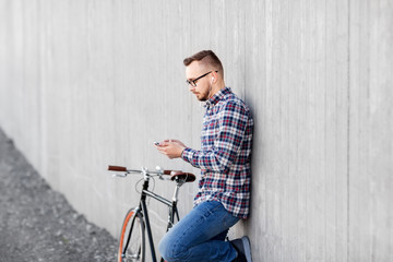 Fototapeta na wymiar technology, leisure and lifestyle- happy young hipster man in earphones with smartphone and fixed gear bike listening to music on city street