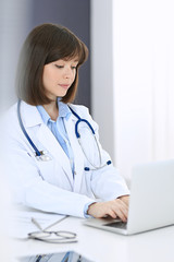 Doctor typing on laptop computer while sitting at the white table in hospital office. Physician at work. Medicine and healthcare concept