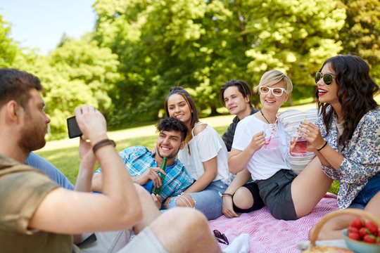friendship, leisure and technology concept - guy taking picture on smartphone of friends drinking non alcoholic drinks at picnic in summer park