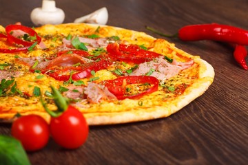 Close up Italian pizza with ham, cheese, pepper and herbs on a brown table decorated by mushrooms, red sweet pepper and cherry tomatoes