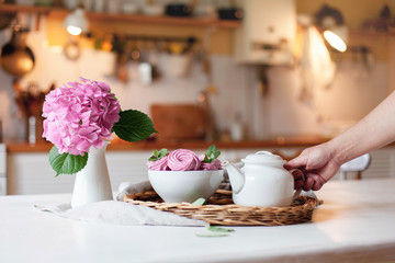 Obraz na płótnie Canvas Woman takes white teapot from serving tray with pink marshmallows. Hydrangea flower is on kitchen table. Morning breakfast with sweets. Tea time and party in cozy home atmosphere hygge.