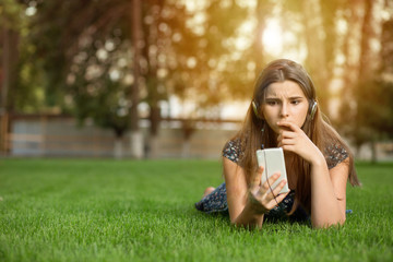 Portrait upset sad skeptical unhappy serious teenager woman looking at phone outdoors lying down on green lawn headphones on head. Multicultural model, mixed race, Asian Russian girl. Copy space.