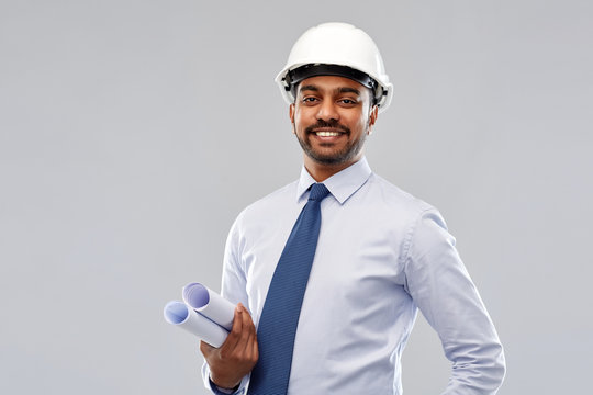 architecture, construction business and building - smiling indian architect or businessman in helmet with blueprints over grey background