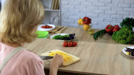 Mature woman cutting fresh yellow pepper on chopping board, salad ingredients