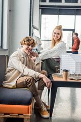 smiling female and male architects with coffee to go working on house model in loft office