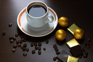 Cup of coffee with beans scattered around and chocolate bars, sweets and candy in golden wrappers on brown wooden business desk, close up, copy space, from above