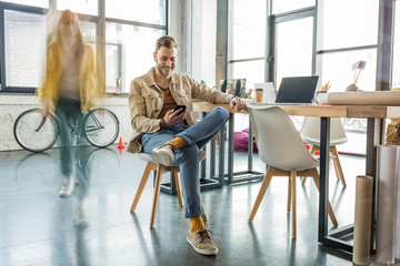 male architect sitting and using smartphone with colleague in motion blur on background in loft office