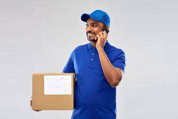 Fototapeta na wymiar mail service, communication and shipment concept - happy indian delivery man with smartphone and parcel box in blue uniform over grey background
