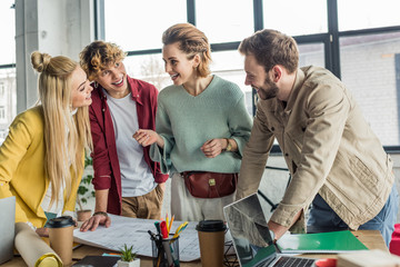 happy group of female and male architects working on blueprint at desk in loft office