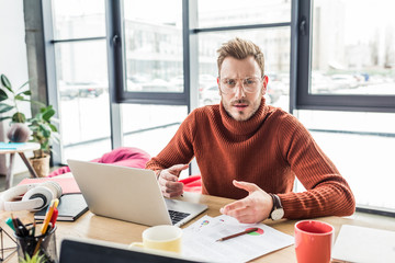 handsome casual businessman sitting at computer desk, looking at camera and working on document in loft office