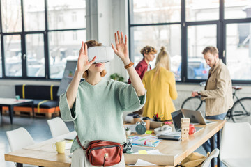 female designer gesturing with hands while having virtual reality experience in loft office with colleagues on background