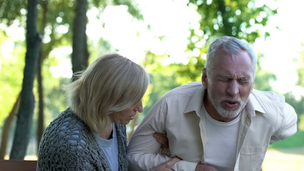 Obraz premium Senior man suffering sharp back pain, walking in park, caring wife helping him