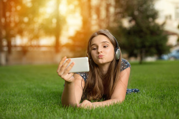 A woman making a grimace blowing a kiss video chatting with friend, looking to  smartphone, girl making duck face outdoors lying down on green lawn headphones on head. Multicultural model, mixed race.