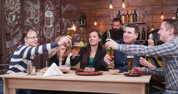 Happy group of friends drinking beer and eating pizza in stylish vintage pub or restaurant. A bearded hipster bartender is working in the background