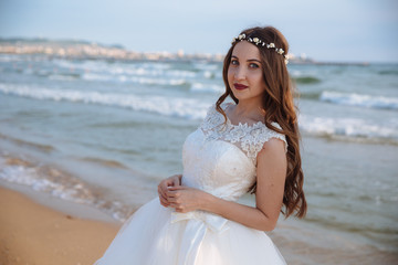 Fototapeta na wymiar Portrait of bride in wedding dress with long curly hair and wreath of flowers on her head at ocean beach